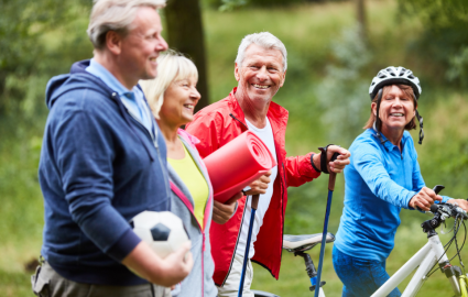 Grupa seniorów na spacerze trzymająca sprzęt sportowy: piłkę, matę, kijki, rower.